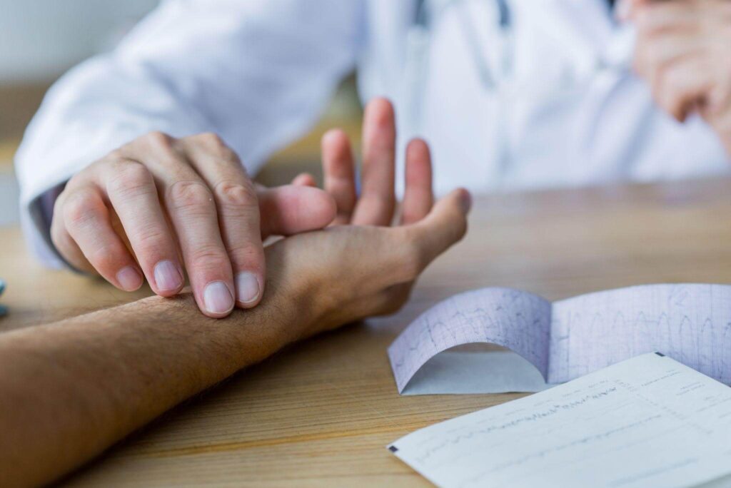 Close das mãos de um médico verificando o pulso de um paciente sobre uma mesa com exames, destacando o cuidado contínuo necessário no controle de doenças autoimunes.