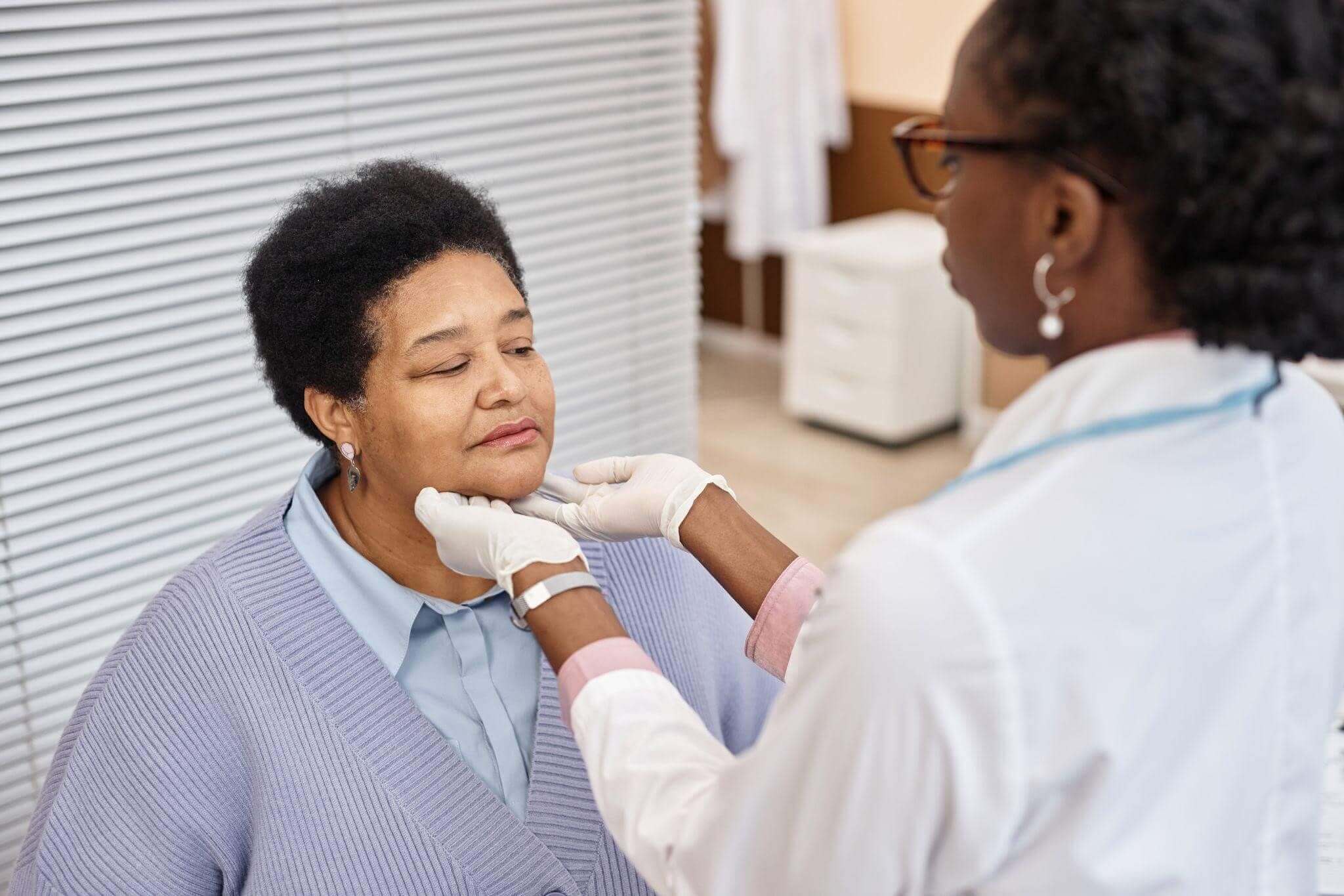 Médica examinando os gânglios linfáticos no pescoço de uma paciente madura durante uma consulta para diagnóstico ou acompanhamento de linfoma. A médica usa luvas brancas e jaleco, enquanto a paciente está sentada e com os olhos fechados.
