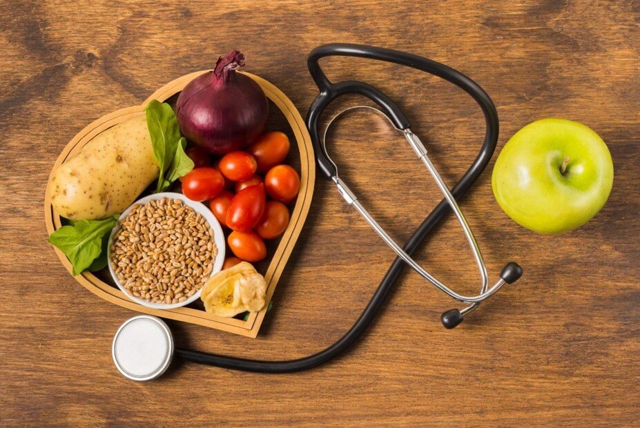 Um estetoscópio ao lado de uma cesta em formato de coração contendo tomate, cebola roxa, batata e grãos integrais. Uma seleção de alimentos naturais focada na prevenção de doenças do coração.